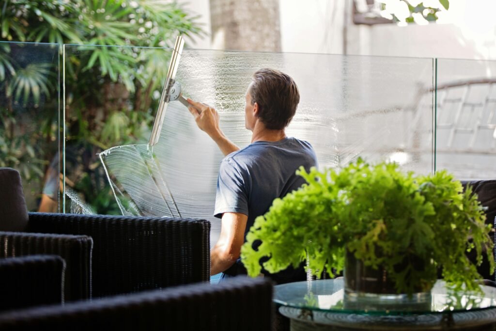 Home A man cleaning a glass barrier outdoors with a squeegee, surrounded by greenery.
